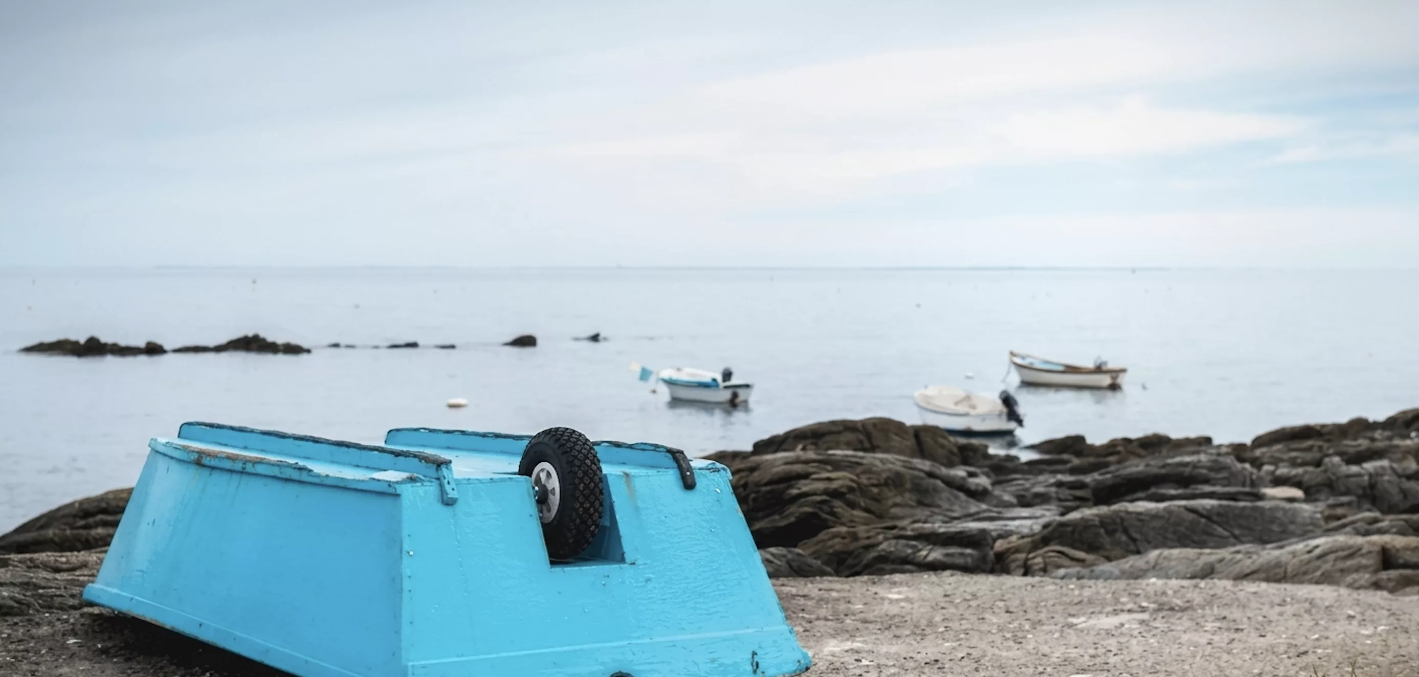 Bateau bleu retourné sur la côte rocheuse de l'Île d'Yeu avec petits bateaux de pêche ancrés dans une mer calme
