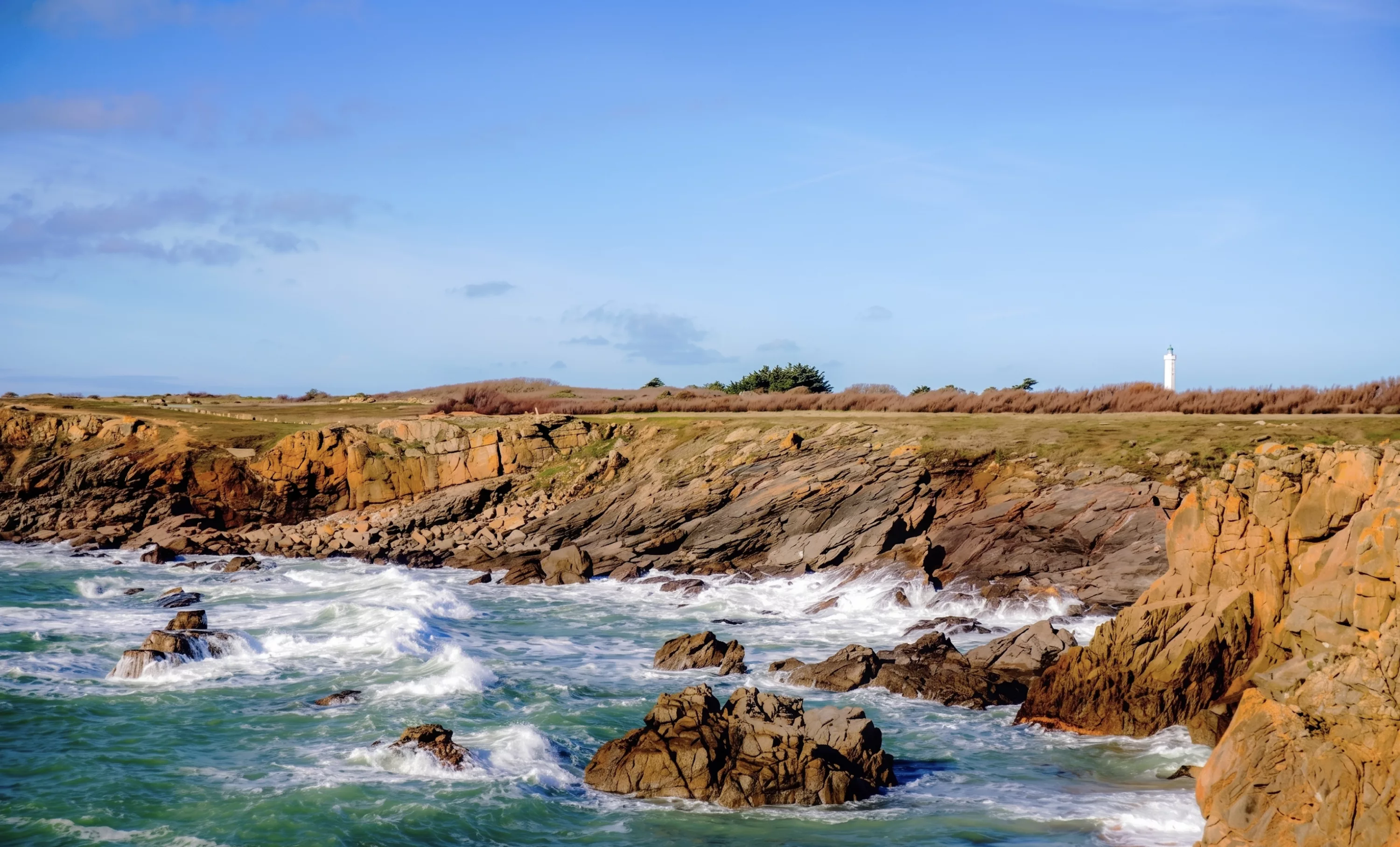 Côte sauvage de l'Île d'Yeu avec rochers de granit ocre battus par les vagues et phare en arrière-plan sous ciel bleu