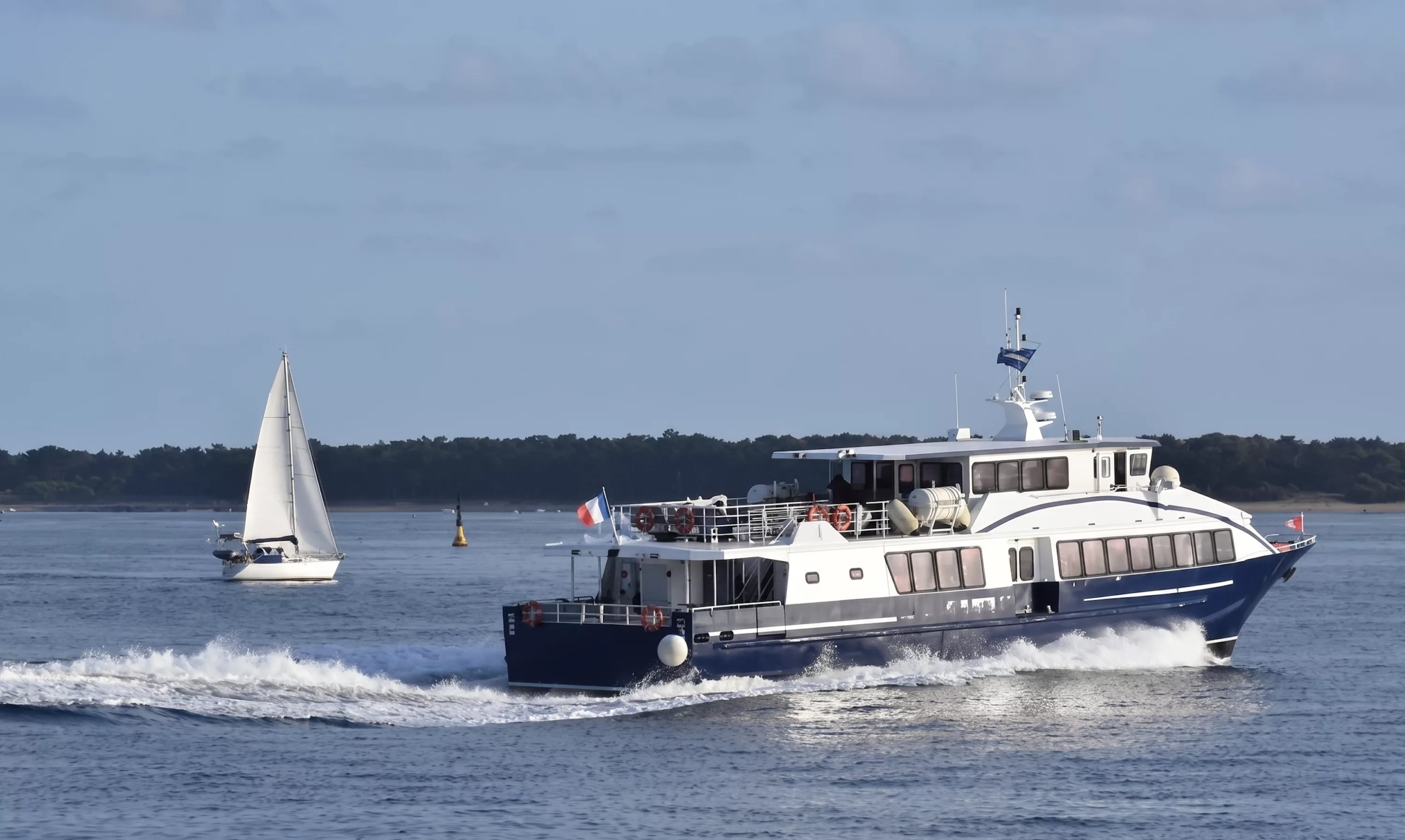 Navette maritime naviguant vers l'Île d'Yeu avec drapeau français, voilier en arrière-plan sur l'océan Atlantique en Vendée