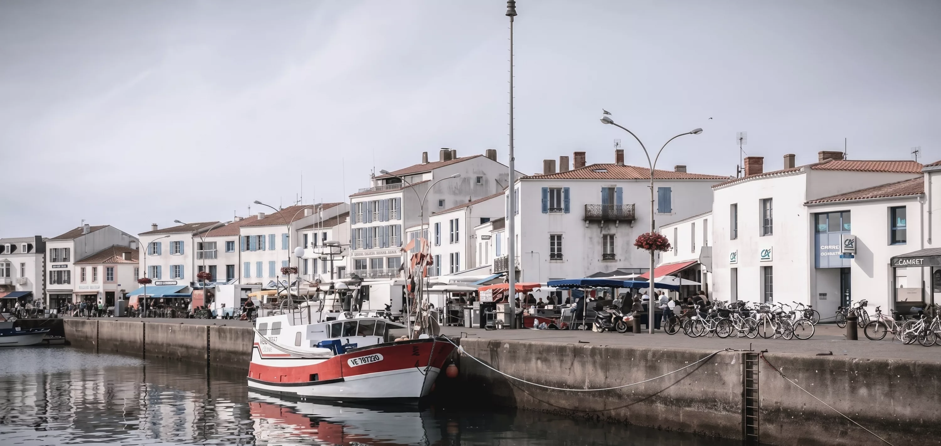 Port de l'Île d'Yeu avec bateau de pêche rouge amarré le long du quai bordé de maisons blanches et commerces animés