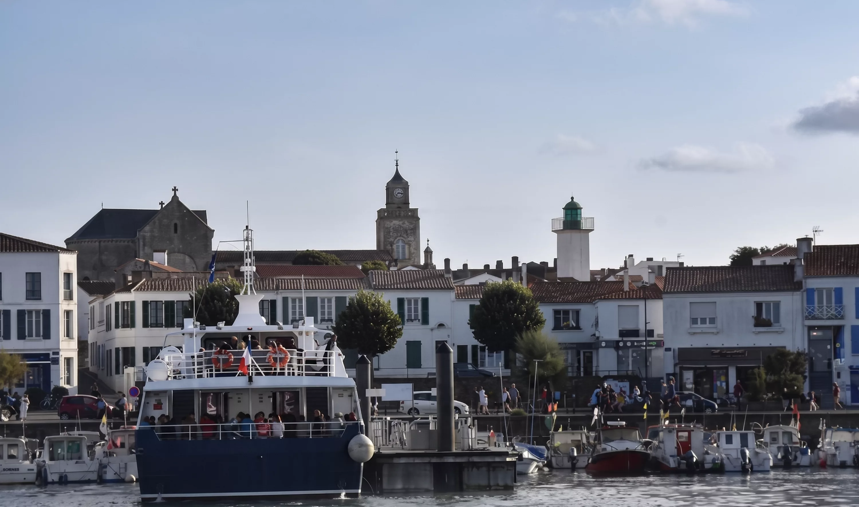 Port-Joinville sur l'Île d'Yeu avec ferry à quai, phare vert et clochers historiques en arrière-plan, Vendée