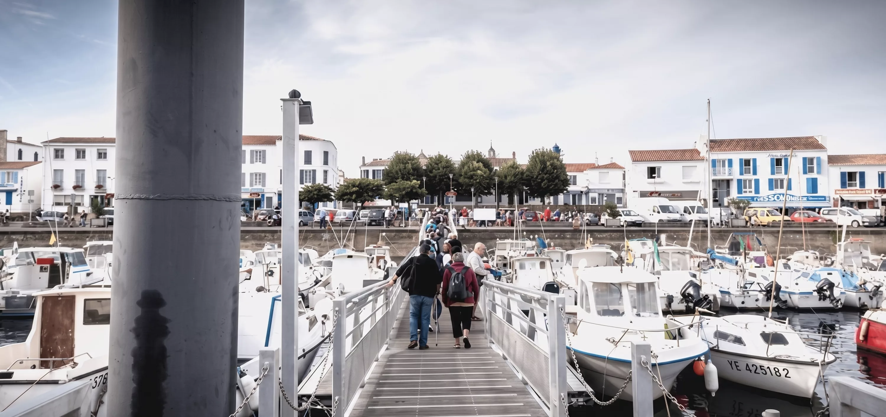 Visiteurs marchant sur la passerelle du port de Port-Joinville avec bateaux amarrés et façades blanches, Île d'Yeu, Vendée