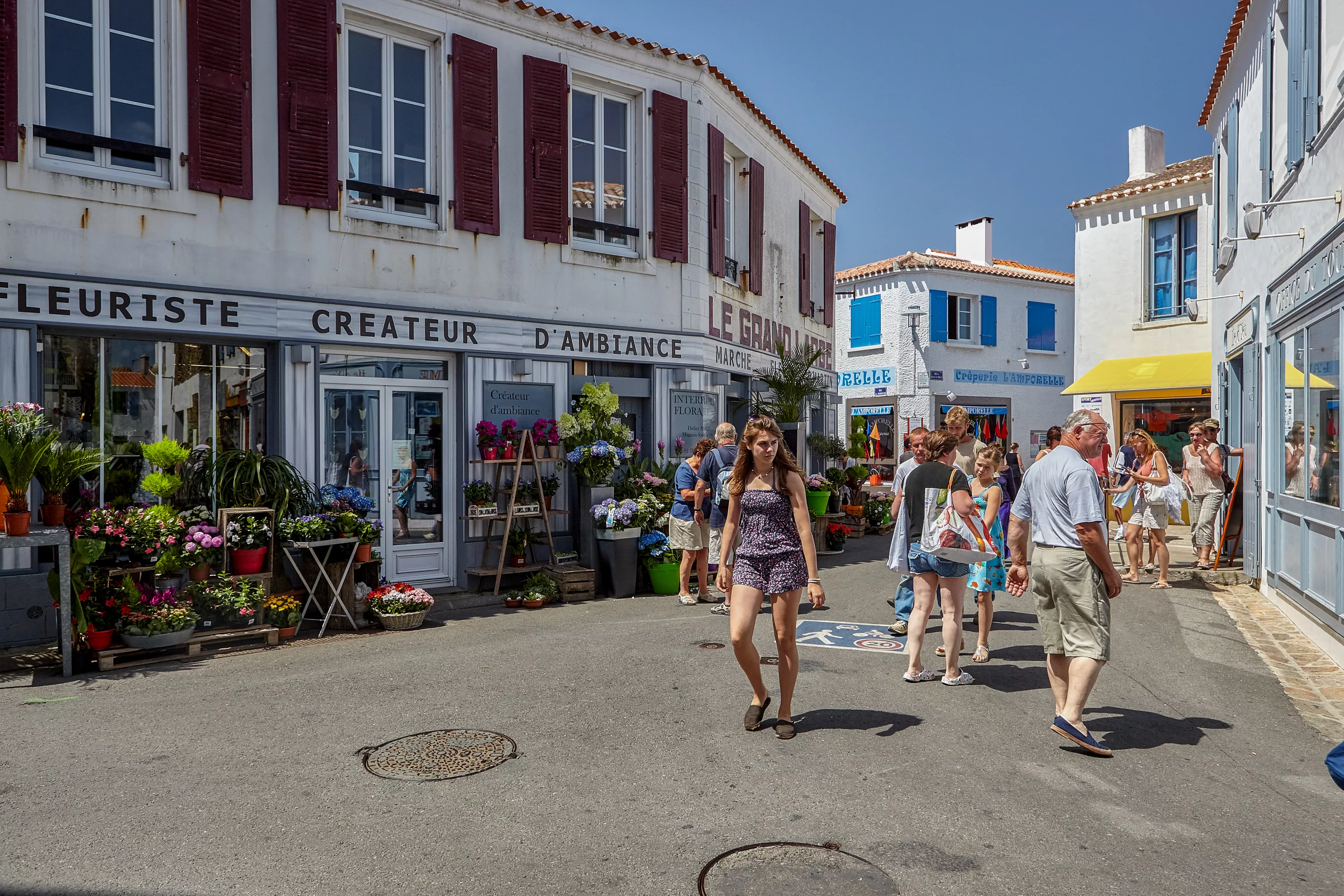 Rue commerçante animée de Port-Joinville sur l'Île d'Yeu avec fleuriste, touristes et façades blanches aux volets colorés